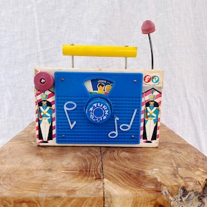 May include: Vintage Fisher-Price toy radio with a blue front panel, yellow handle, and a pink antenna. The radio features a dial that says "Turn" and illustrations of children. The sides are decorated with toy soldier graphics.