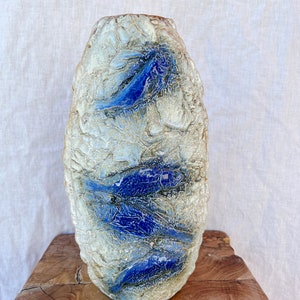 May include: A textured ceramic vase with a light beige and blue color scheme. The vase features a series of blue, fish-like shapes. The vase is displayed on a wooden surface.
