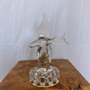 Op de afbeelding: Een heldere glazen duifsculptuur met uitgespreide vleugels. De vogel staat op een ronde, getextureerde basis. De sculptuur wordt tentoongesteld op een houten oppervlak tegen een witte achtergrond.