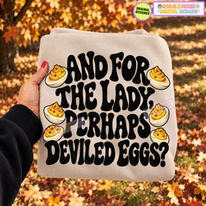 May include: A beige sweatshirt featuring black text that reads, "AND FOR THE LADY, PERHAPS DEVILED EGGS?" The design includes five illustrations of halved deviled eggs with yellow yolks and green garnish scattered around the bold lettering.