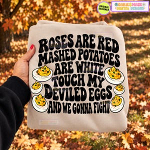 May include: A beige sweatshirt featuring black text that reads: ROSES ARE RED MASHED POTATOES ARE WHITE TOUCH MY DEVILED EGGS AND WE GONNA FIGHT. The design includes illustrations of four halved deviled eggs with yellow yolks and orange paprika.
