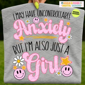 Può includere: Una maglietta grigia con la scritta "I MAY HAVE UNCONTROLLABLE Anxiety BUT I'M ALSO JUST A Girl" in rosa, nero e giallo. Il design include faccine sorridenti, stelle e margherite.