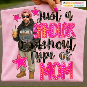 Peut inclure: T-shirt rose avec un graphisme d'un homme portant des lunettes de soleil et un bonnet, tenant un café. Le texte dit "Just a Sandler Crashout Type of MOM" avec des étoiles roses. Le t-shirt est tenu par une main avec du vernis à ongles rose.