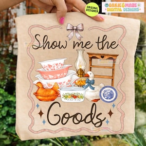Op de afbeelding: Een beige draagtas met een vintage-stijl illustratie. Het ontwerp bevat de tekst "Show me the Goods" en omvat gestapelde kommen, een kippenvormige schaal, een eend, een kleine houten plank en een blauw-wit bord, allemaal omlijst met een roze rand.