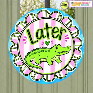 May include: Round door sign with a cartoon alligator and the word "Later" in a playful font. The sign has a pink and white striped background, a scalloped edge, and a blue border. A green heart is between the words.