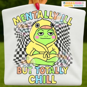 Puede incluir: Camiseta blanca con un diseño de una rana de dibujos animados con un disfraz de pato amarillo y el texto "MENTALLY ILL BUT TOTALLY CHILL" en letras coloridas. El diseño incluye un patrón de bandera a cuadros en el fondo.