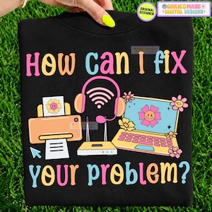 May include: A black t-shirt featuring the text How Can I Fix Your Problem? in colorful letters. The design includes a peach-colored printer, a pink headset with a Wi-Fi symbol, and a yellow laptop, all decorated with small pink and yellow flowers.