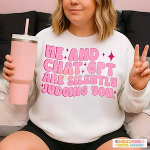 Könnte beinhalten: Ein weißes Sweatshirt mit dem Text "ME AND CHAT GPT ARE SILENTLY JUDGING YOU" in rosa Buchstaben. Ein rosa Becher mit weißem Deckel und Strohhalm wird in der linken Hand gehalten. Die Person macht auch ein Friedenszeichen.
