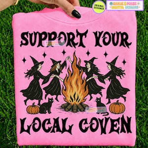 May include: A bright pink sweatshirt featuring a black graphic of four witches in pointed hats dancing around a large orange and yellow bonfire. Two black cats and two orange pumpkins are at the base. The text reads SUPPORT YOUR LOCAL COVEN.