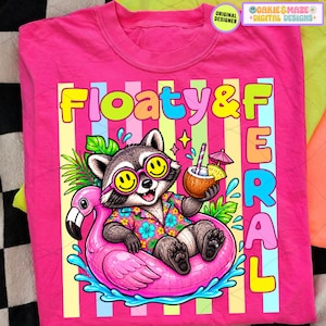 Op de afbeelding: Een felroze t-shirt met een cartoon wasbeer met een zonnebril en een Hawaiiaans shirt, drijvend op een roze flamingo zwemband. Het shirt heeft de woorden "Floaty & Feral" in kleurrijke letters.