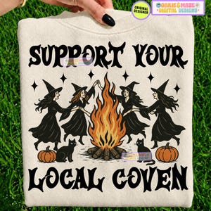 May include: A cream-colored sweatshirt featuring a black graphic print of four witches dancing around a central bonfire. The text reads SUPPORT YOUR LOCAL COVEN in a stylized black font. Two black cats and two orange pumpkins are at the base.