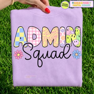 May include: A light purple crewneck sweatshirt featuring the text ADMIN in large, multi-patterned block letters and the word Squad in black cursive script. The design includes a pink flower on the left and a blue flower on the right side.
