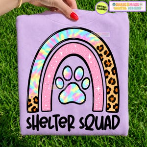 May include: A light purple sweatshirt featuring a graphic of a rainbow with a paw print in the centre. The rainbow has tie-dye, glittery pink, and leopard print sections. The text "SHELTER SQUAD" is printed in black below the rainbow design.