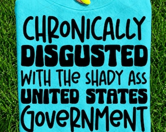 May include: A bright turquoise crewneck sweatshirt featuring bold black lettering that reads: CHRONICALLY DISGUSTED WITH THE SHADY ASS UNITED STATES GOVERNMENT. The text is arranged in four lines with a playful, rounded font style on the front of the garment.