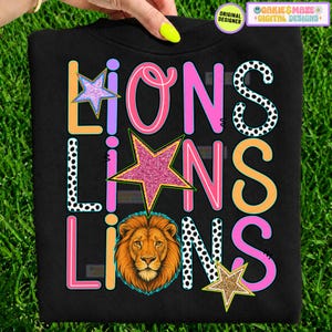 May include: A black sweatshirt featuring the word LIONS repeated three times in a graphic design. The letters are rendered in various colors including orange, pink, and black-and-white polka dots, accented by three glittery stars and a lion head.