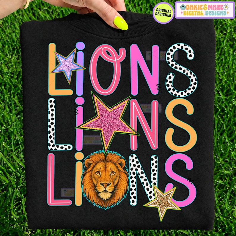 May include: A black sweatshirt featuring the word LIONS repeated three times in a graphic design. The letters are rendered in various colors including orange, pink, and black-and-white polka dots, accented by three glittery stars and a lion head.