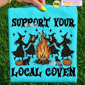 May include: A bright turquoise sweatshirt featuring a black graphic of four witches dancing around a campfire. The text reads SUPPORT YOUR LOCAL COVEN in a bold black font. Two black cats and two orange pumpkins are positioned at the base.