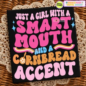 Puede incluir: Un estampado gráfico rectangular negro con el texto rosa JUST A GIRL WITH A, SMART MOUTH y ACCENT. La palabra AND A es azul claro y CORNBREAD es naranja. El diseño incluye pequeñas estrellas azul claro y un pequeño icono de flor naranja.
