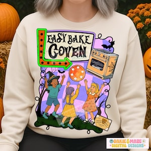 May include: Cream-colored sweatshirt featuring a Halloween-themed graphic. The design includes the text "Easy Bake Coven" above an illustration of three children, a pie, an oven, and a Ouija board. The graphic is outlined with black lines.