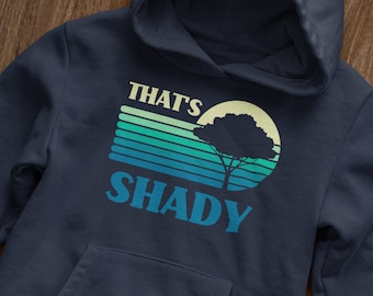May include: A navy blue hoodie with a retro-style graphic design featuring a tree silhouette in front of a sunset with the text "THAT'S SHADY" in a blue and yellow font.