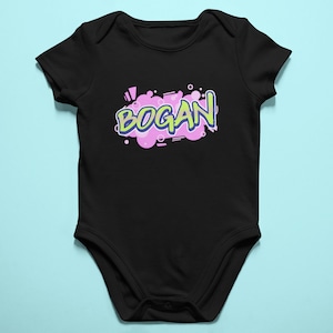 May include: Black baby bodysuit with the word "BOGAN" in a graffiti-style design. The text is in green and outlined in blue, set against a pink and purple cloud-like shape. The bodysuit is made of soft fabric.