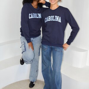 Puede incluir: Dos personas vestidas con sudaderas azul marino con la palabra "CAROLINA" impresa en blanco. Tambi&eacute;n llevan vaqueros azules. Una persona lleva vaqueros cargo azul claro y zapatos negros. La otra persona lleva vaqueros azules acampanados.