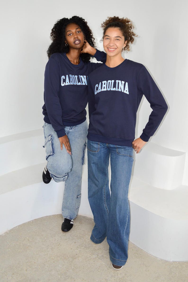 Puede incluir: Dos personas vestidas con sudaderas azul marino con la palabra "CAROLINA" impresa en blanco. Tambi&eacute;n llevan vaqueros azules. Una persona lleva vaqueros cargo azul claro y zapatos negros. La otra persona lleva vaqueros azules acampanados.