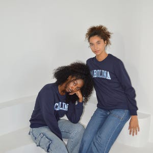Puede incluir: Dos personas vestidas con camisetas de manga larga azul marino con la palabra "CAROLINA" impresa en azul claro. Tambi&eacute;n llevan vaqueros azules. El fondo es una pared blanca lisa.