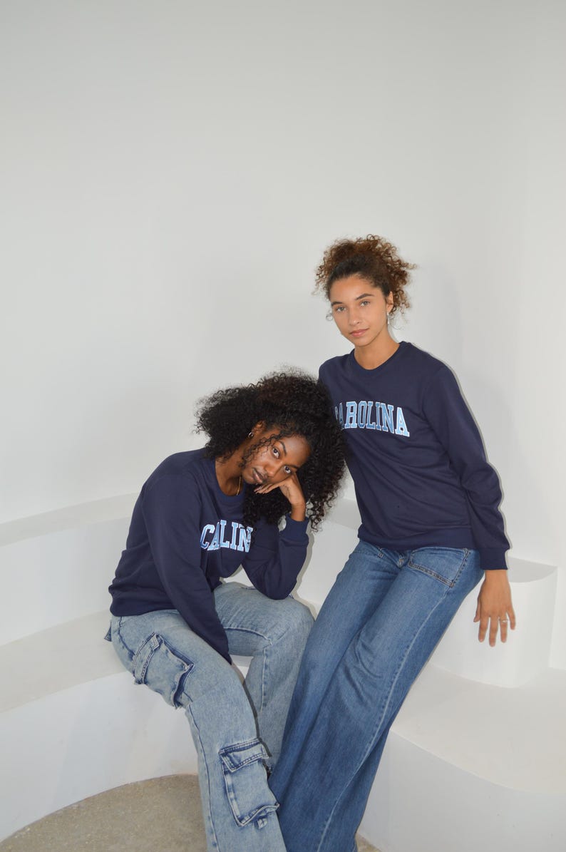 Puede incluir: Dos personas vestidas con camisetas de manga larga azul marino con la palabra "CAROLINA" impresa en azul claro. Tambi&eacute;n llevan vaqueros azules. El fondo es una pared blanca lisa.