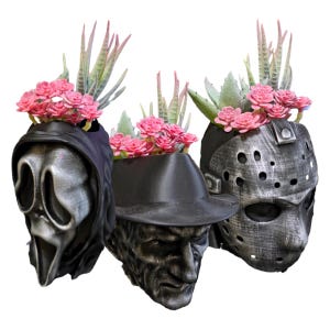 Peut inclure: Trois pots décoratifs en forme de masques et de visages de films d'horreur. Les pots sont peints dans des tons argent et noir, et chacun contient des fleurs roses et des succulentes. Le masque de Scream, le visage de Freddy Krueger et le masque de Jason Voorhees sont présentés.