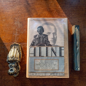 May include: A hardcover book titled Aline by Carole Klein, featuring a portrait of Aline Bernstein on the cover. Beside the book is a brass door knocker with a decorative leaf design and a long, narrow, dark metal object with vertical engravings.