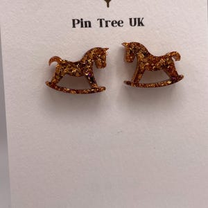 May include: A pair of gold-coloured rocking horse earrings with glitter detailing. The earrings are presented on a white card, with the text "Pin Tree UK" and "Handmade With Love".