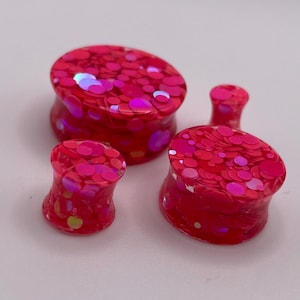 May include: Four red and pink ear gauges with a glittery, speckled design. The plugs are flared and have a rounded, oval shape. The gauges are made of a translucent material.