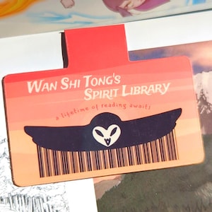 Puede incluir: Un marcapáginas rojo y naranja con el texto "Wan Shi Tong's Spirit Library" y un gráfico de un búho. El marcapáginas tiene la frase "a lifetime of reading awaits". El marcapáginas está encima de un libro y un dibujo.