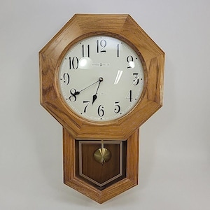 May include: An octagonal wall clock with a light brown oak wood frame and a white face featuring black Arabic numerals. The Howard Miller brand name is printed on the dial. A brass-colored pendulum hangs in the lower wooden section.