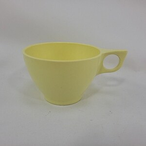 May include: A pale yellow cup with a smooth, matte finish and a wide, circular handle integrated into the side. The cup features a tapered body that narrows toward the base, providing a simple, functional design for holding liquids or small items.