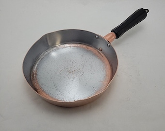May include: A copper-colored frying pan with a black handle. The pan has a silver interior and a copper exterior. The handle is black and angled. The pan is round and has a small lip on one side.