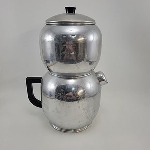 May include: A vintage, two-part, aluminum coffee percolator with a black handle and lid knob. The percolator has a shiny, reflective surface and a spout for pouring. The top section has a clear water level indicator.