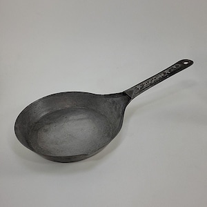 May include: A vintage, oval-shaped, metal pan with a long handle. The pan has a dark, matte finish and a slightly textured surface. The handle is stamped with the word "DeLuxe" and star designs. The pan is designed for cooking.