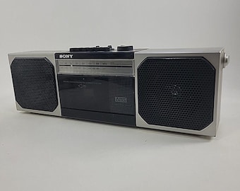 Op de afbeelding: Een zilverkleurige en zwarte draagbare Sony stereo-cassettespeler met twee geïntegreerde luidsprekers aan de zijkanten. De voorzijde bevat een radiofrequentiedisplay, een centraal cassettecompartiment en een klein AMS-logo. Zwarte knoppen aan de bovenkant.