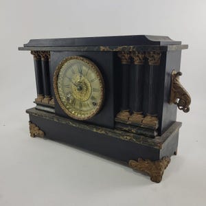 May include: A rectangular mantel clock featuring a black finish with faux marble accents. The clock face is circular with gold-toned filigree, black Roman numerals, and ornate gold-toned metal feet and side handles. Four fluted columns flank the dial.