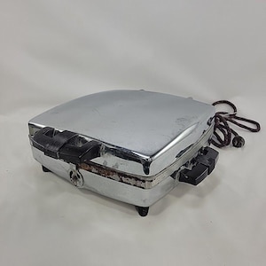 May include: A vintage chrome waffle maker with a square, polished metal exterior and black plastic handles on the sides. The appliance features a hinged lid, a front latch mechanism, and a dark, braided electrical cord with a plug attached.