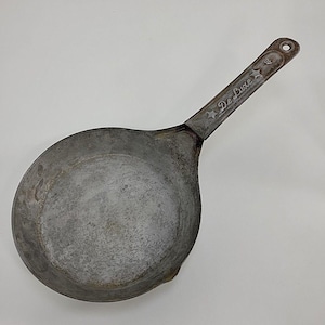 May include: A vintage, well-worn metal ladle with a long handle. The handle is marked "De Luxe" with star accents. The ladle has a rounded bowl and a small spout. The metal has a mottled, aged appearance.
