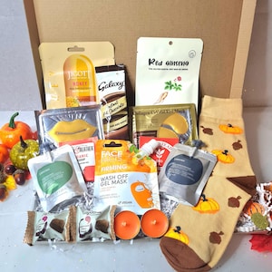 Op de afbeelding: Een geschenkdoos gevuld met schoonheidsproducten en lekkernijen. Bevat gezichtsmaskers, oogmaskers, chocolade en sokken met pompoenthema. De doos bevat ook twee oranje kaarsen en decoratief herfstblad.