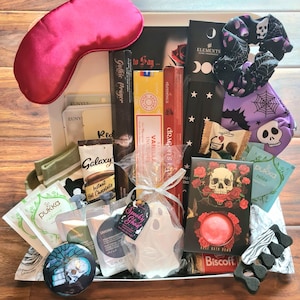 Op de afbeelding: Een thematische geschenkdoos met een roze slaapmasker, wierook, een badbruisbal, sokken, thee en een spookvormige zeep. De doos heeft een griezelig thema met schedel- en rozenafbeeldingen.