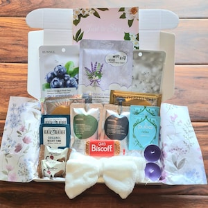 Op de afbeelding: Een witte geschenkdoos met huidverzorgingsproducten, een witte strik, kaarsen en een Biscoff-koekje. De doos bevat gezichtsmaskers, lotions en een kaart met de tekst "Just to Say".
