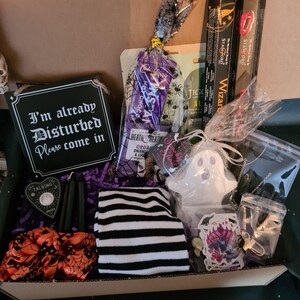 May include: A Halloween gift box containing a variety of items. The box includes a sign saying "I'm already Disturbed Please come in", black candles, a ghost-shaped item, striped socks, and incense sticks. The box is decorated with purple paper and orange and black accents.