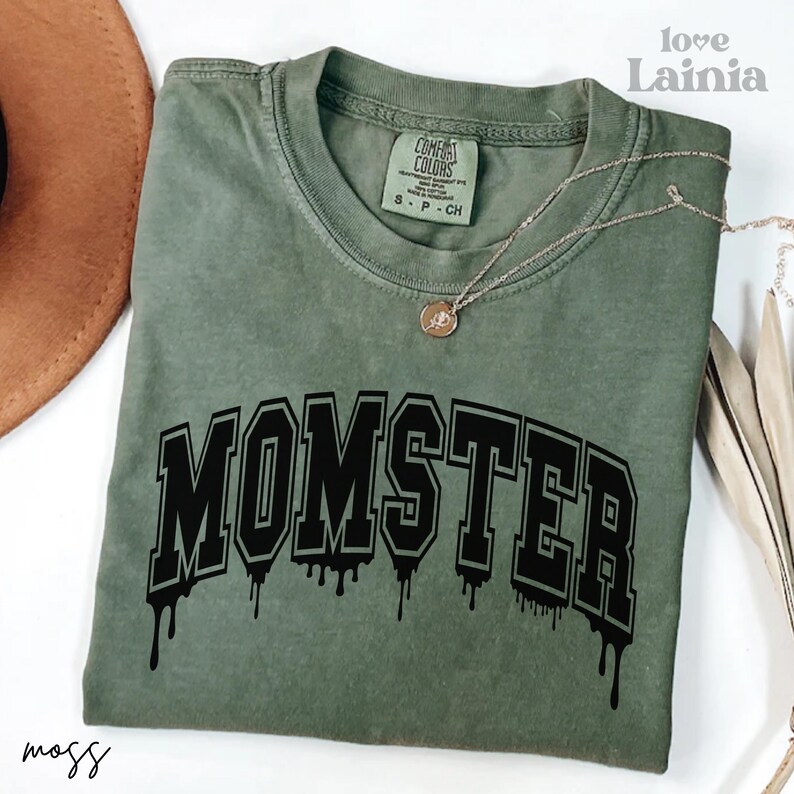 May include: An olive green short-sleeve t-shirt with a crew neckline. The shirt features the word MOMSTER in black collegiate-style lettering with a dripping effect at the bottom. A gold-toned necklace with a small circular pendant rests on the shirt.