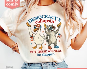 May include: Cream-colored t-shirt with a humorous graphic. The design features a cartoon duck and raccoon holding hot dogs, with text reading "DEMOCRACY'S collapsin' BUT THESE WIENERS be slappin'". The shirt has a "soft & comfy" label.
