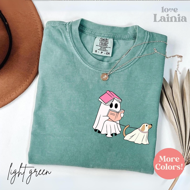 Op de afbeelding: Lichtgroen Comfort Colors T-shirt met een afbeelding van een spook dat een boek leest en een hond uitlaat, beide in spookkostuums. Het shirt heeft een ronde hals en korte mouwen. Een zilveren ketting met een hanger ligt op het shirt.